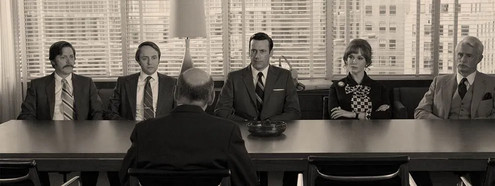 Vijf mensen zitten op een rij tegenover een gespreksleider aan een vergadertafel, sepia still uit Mad Men