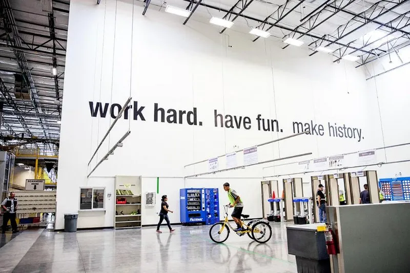 Bedrijfshal met op de muur de tekst work hard, have fun, make history en een medewerker op een fiets
