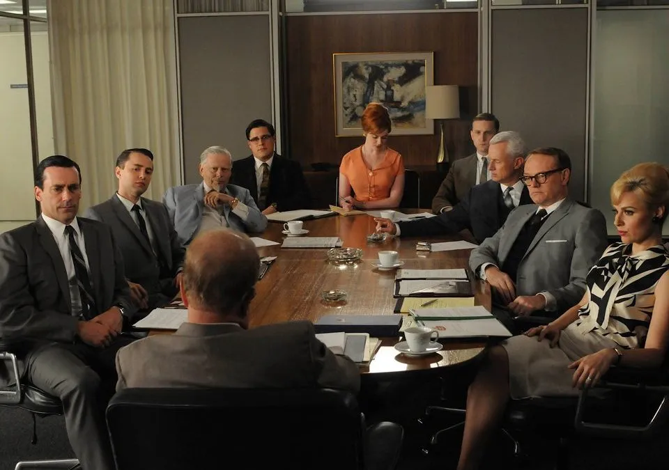 Vergadering rond een grote ovalen tafel op een kantoor uit de jaren zestig, still uit Mad Men