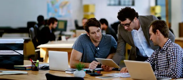 Drie collega's overleggen samen achter een tablet en laptops op een open kantoor